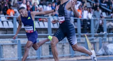 Two male athletes in action sprinting across the finish line with the crowd in a stadium in the background.