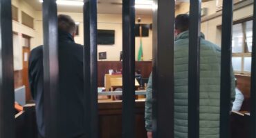 Two men photographed from the back behind bars in a courtroom.