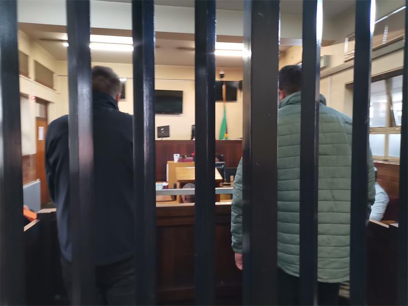 Two men photographed from the back behind bars in a courtroom.