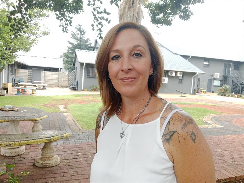 A white woman with shoulder-length brown hair wearing a white sleeveless shirt with tattoos on her arms with a building in the background.