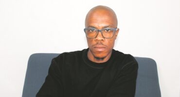 A black male with a shaved head wearing black rimmed glasses and a black shirt.