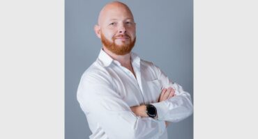 A white man with a bald head and red beard and moustache wearing a white shirt and standing with his arms crossed.