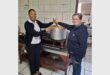 Two women, one black and one white, standing at a stove stirring a big steel pot of soup.