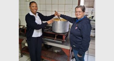 Two women, one black and one white, standing at a stove stirring a big steel pot of soup.