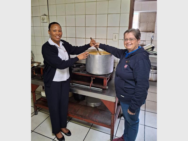 Two women, one black and one white, standing at a stove stirring a big steel pot of soup.