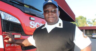 A black male wearing glasses and a cap, white shirt and black pullover next to the front of a fire truck.