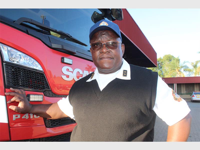 A black male wearing glasses and a cap, white shirt and black pullover next to the front of a fire truck.
