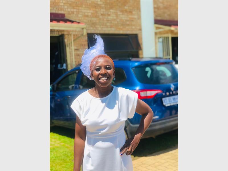 A young black woman standing with her arm on her side smiling at the camera, wearing a white dress with a dark coloured car in the background.