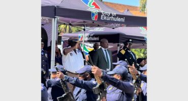 People standing under a City of Ekurhuleni gazebo while a parade of police officers comes past.