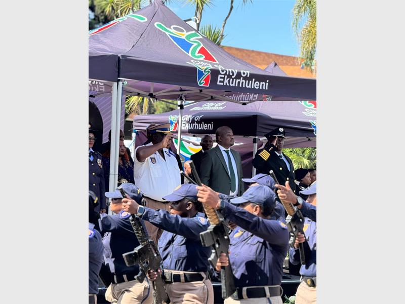 People standing under a City of Ekurhuleni gazebo while a parade of police officers comes past.