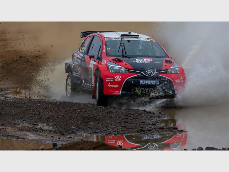 A black and red rally car racing through water and mud, making a big splash. 