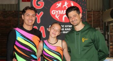 A male and female gymnast in brightly coloured shirts with their coach wearing a springbok jacket.