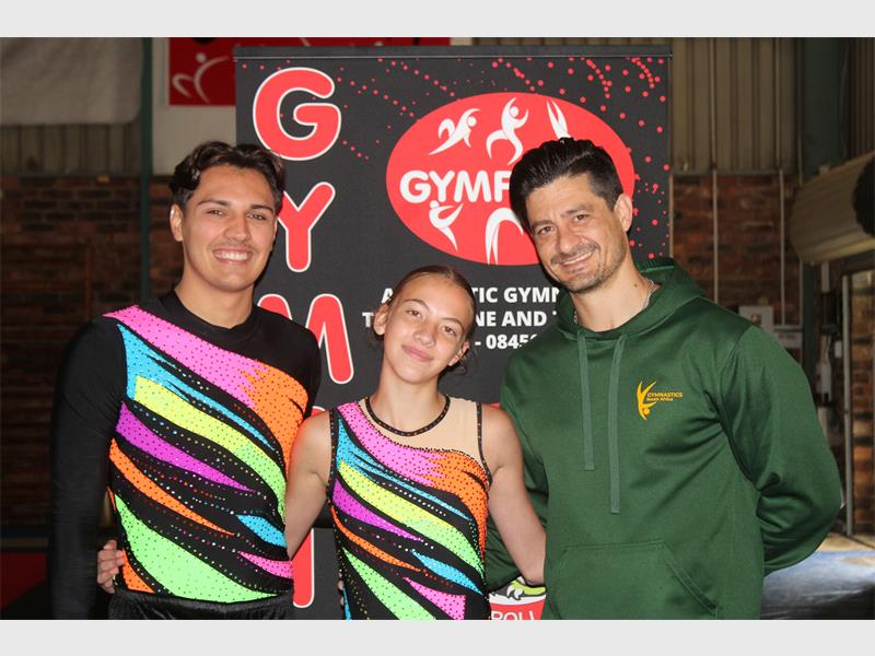 A male and female gymnast in brightly coloured shirts with their coach wearing a springbok jacket.