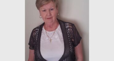 A white woman with short hair, wearing a white shirt with a short sleeved black knitted top over it.