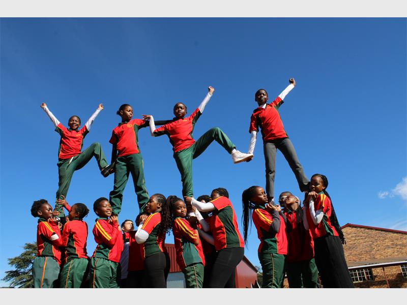 Drum majorettes performing a set with four standing on the others' shoulders, all dressed in red shirts and green plants. 