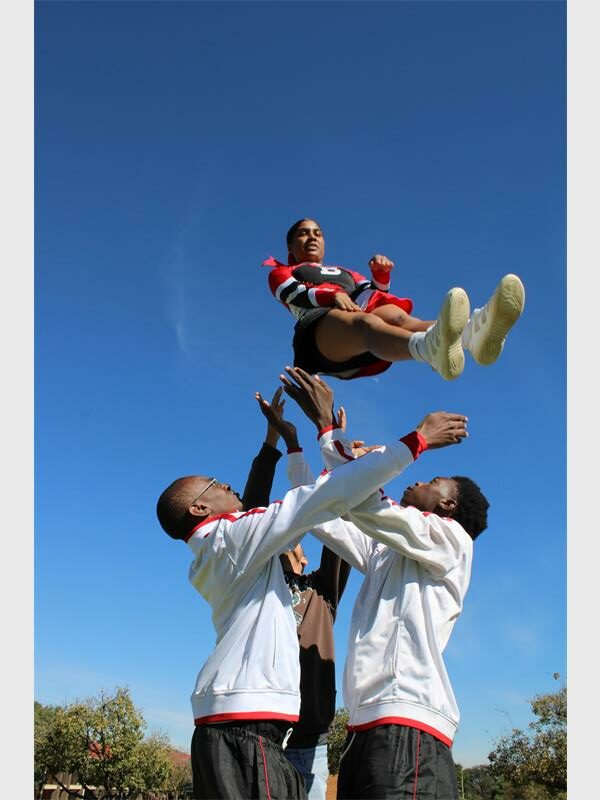 Two male learners with arms outstretched upwards as they fling a female drum majorette into the air and wait to catch her. 