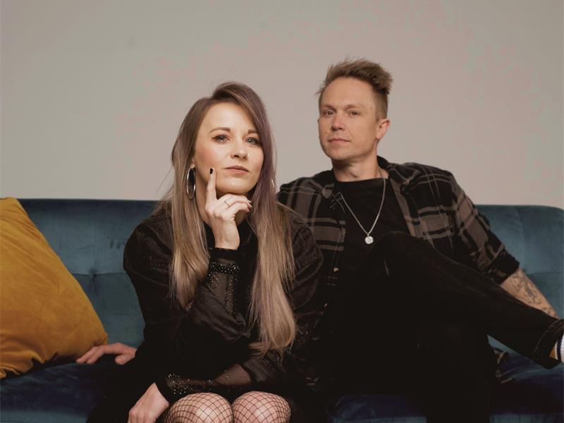 A young white woman and man sitting ona blue couch wearing black. She has long blonde hair.