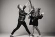 A black and white image of a man and a woman dancing and reaching up towards a guitar she is holding.