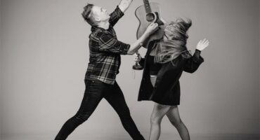 A black and white image of a man and a woman dancing and reaching up towards a guitar she is holding.