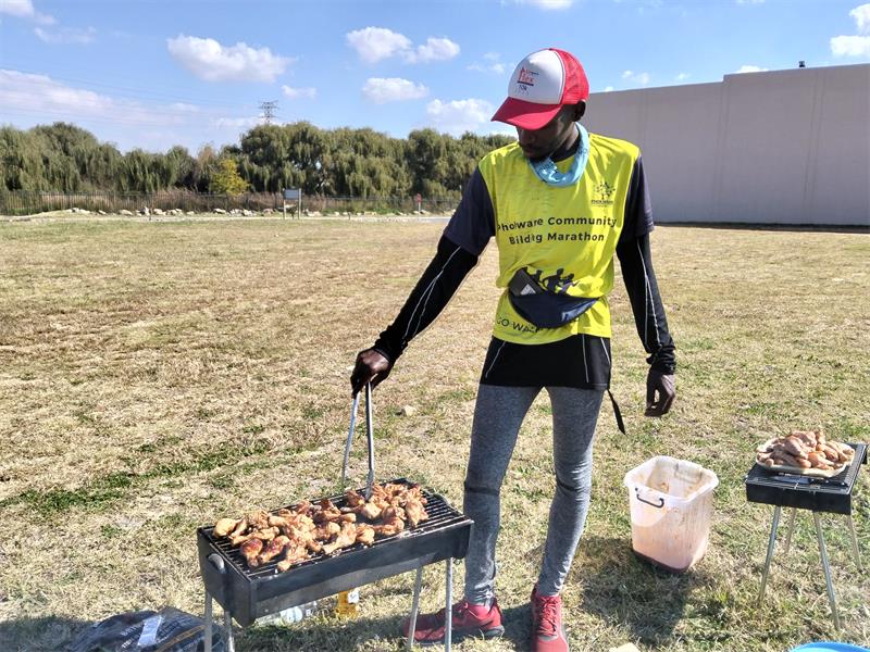 A black man with a bright yellow vest and athletic clothes is cooking chicken drumsticks on a small braai on a large open grass lawn. To the right another braai with more chicken is visible, as well as a white bucket.