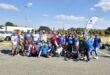 A large group of athletes in activewear in a parking lot outside a Planet Fitness gym. Behind and to the right of the group is a banner with the City of Ekurhuleni logo and some text on.