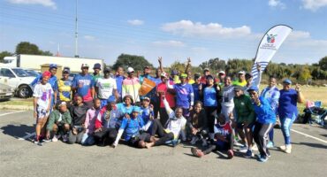 A large group of athletes in activewear in a parking lot outside a Planet Fitness gym. Behind and to the right of the group is a banner with the City of Ekurhuleni logo and some text on.