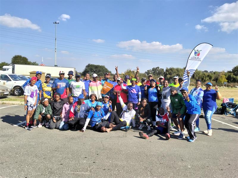 A large group of athletes in activewear in a parking lot outside a Planet Fitness gym. Behind and to the right of the group is a banner with the City of Ekurhuleni logo and some text on.