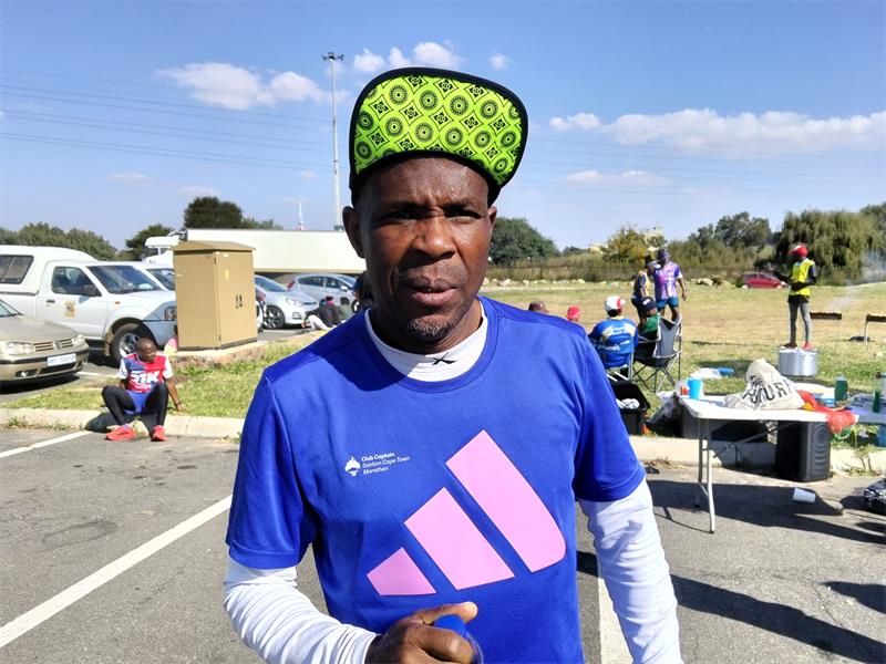 A black man in athletic clothes with a visor that is flipped up. Behind him is a field with some people, two braais, and some cars visible.