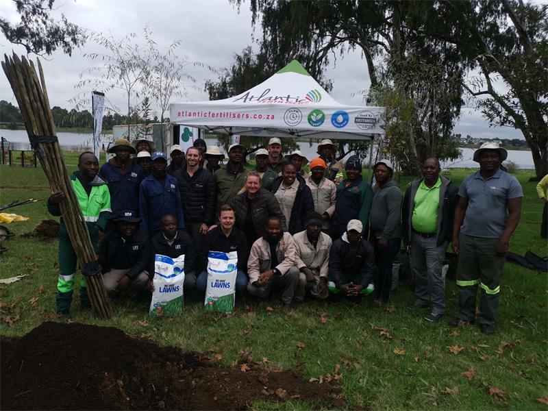 A big group of people posing with bags of soil for tree planting at Homestead Dam with a white gazebo in the background.