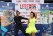 A black couple, the man dressed all in black and the woman in a flowing yellow dress performing a dance move in front of a banner reading Legends are Made.