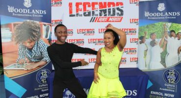 A black couple, the man dressed all in black and the woman in a flowing yellow dress performing a dance move in front of a banner reading Legends are Made.