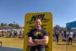 A white male with his arms crossed in front of a yellow banner, wearing a black T-shirt.