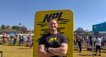 A white male with his arms crossed in front of a yellow banner, wearing a black T-shirt.