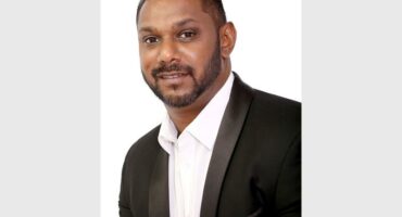 An Indian male with short neat hair, a beard and moustache and dressed in a white shirt and black jacket.