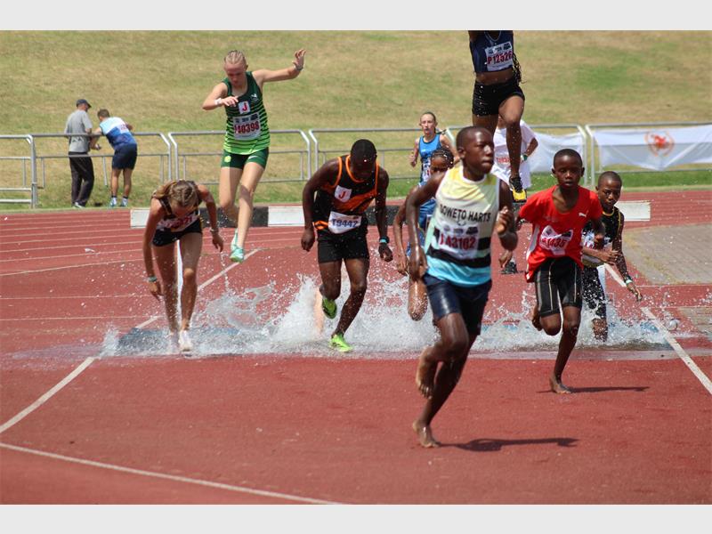 A group of female and male athletes taking part in a steeple chase through water on an athletics track.