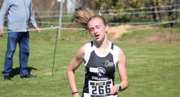 A young female athlete with her blonde ponytail blowing in the wind pictured during a race, wearing blue and white kit.