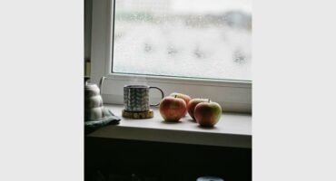 Cups and fruit on a cupboard in front of a window that depicts moisture drops on the outside.