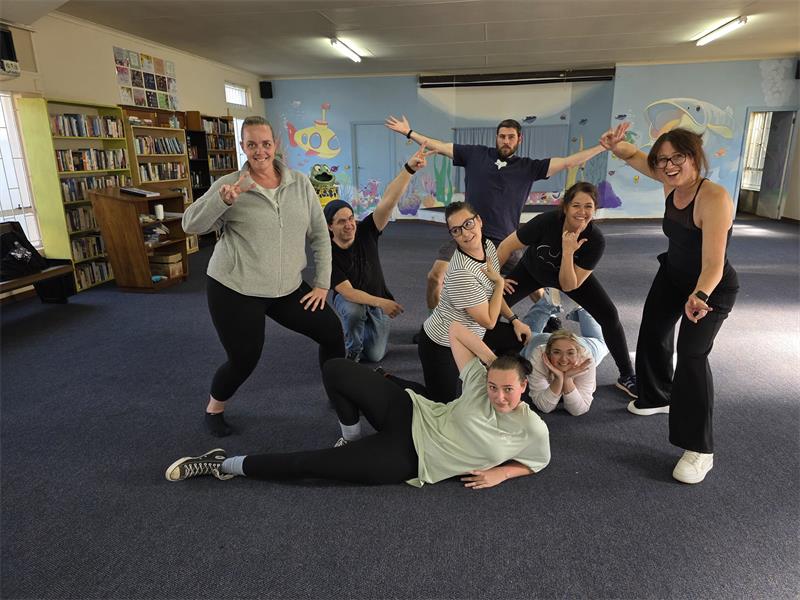 A group of theatre performers wearing black pants and T-shirts in carpeted room in various poses for the camera to promote their latest production.