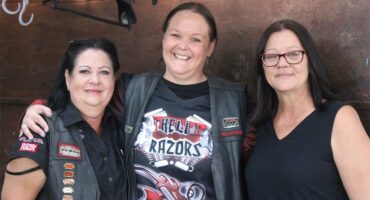 Three female members of Hell Razors biker club standing next to each other smiling for the camera, all dressed in the club's shirts and jackets.