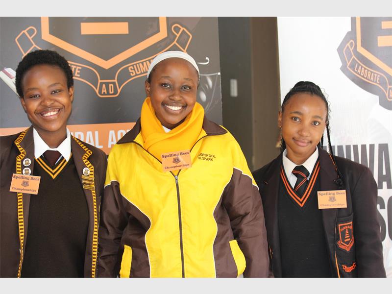 Three black female learners in brown and yellow uniform standing in a row in front of the school badge displayed on a wall. 