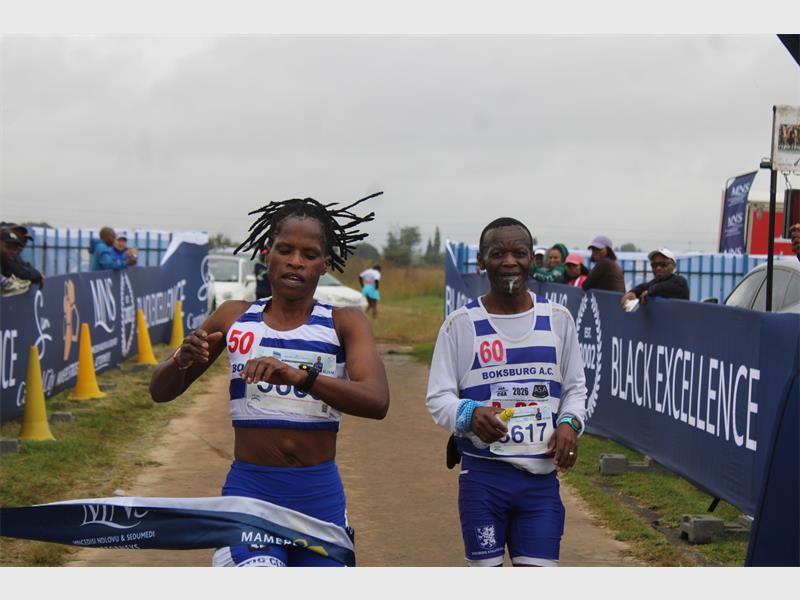Two runners dressed in blue and white kit, a female and a male, coming over the finish line at a marathon.