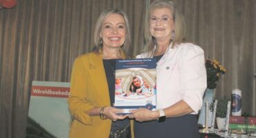 Two white women, one wearing a yellow jacket and black dress and the other a white shirt, holding a book and smiling for the camera.