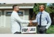 Two males shaking hands while facing each other dressed in suits with a house in the background.
