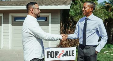 Two males shaking hands while facing each other dressed in suits with a house in the background.