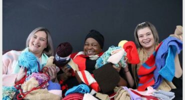 A black female wearing a black beanie flanked by two white women, all draped in knitted scarves and holding balls of wool.
