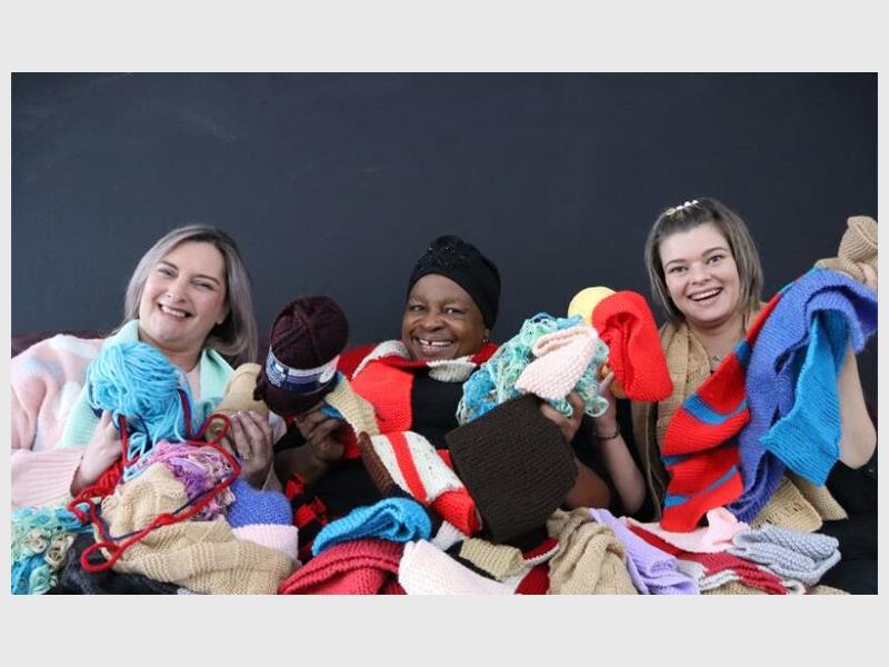 A black female wearing a black beanie flanked by two white women, all draped in knitted scarves and holding balls of wool.