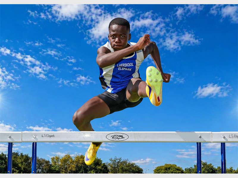 A young black athlete wearing blue and white kit jumping over a hurdle on an athletic track.