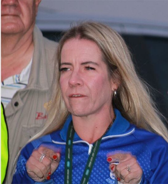A white female with long blonde hair wearing a blue anorak and holding her hands up to her chest.