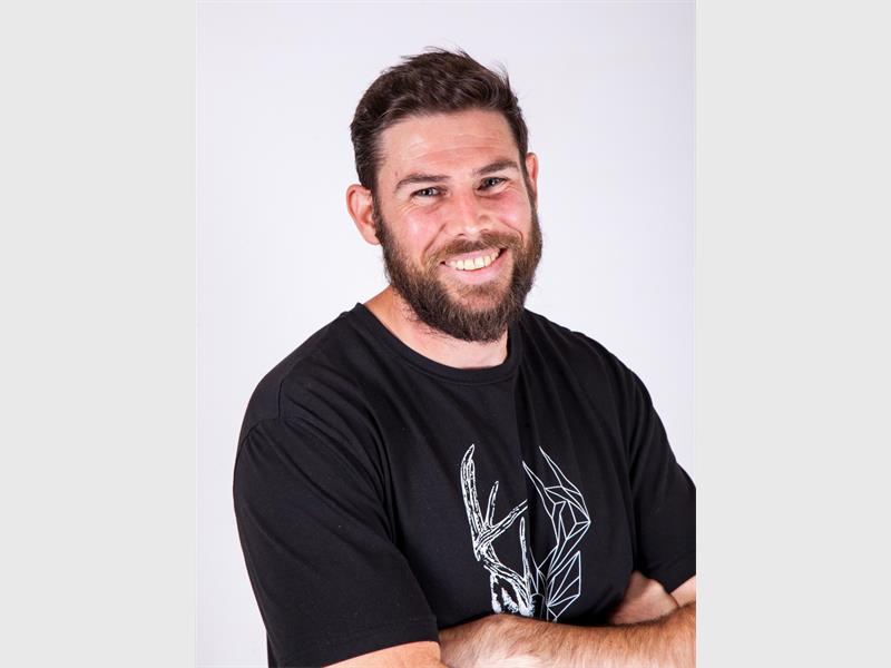 A young white man with short dark hair and a beard and moustache wearing a black T-shirt and with his arms cross over his chest, smiling for the camera.