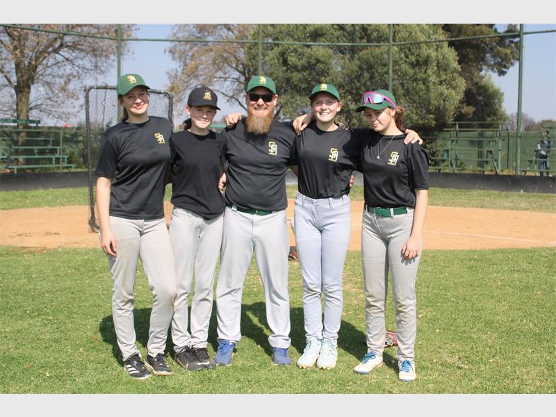 A white male with a beard flanked by four young white female baseball players, all dressed in black shirts, white pants and caps.